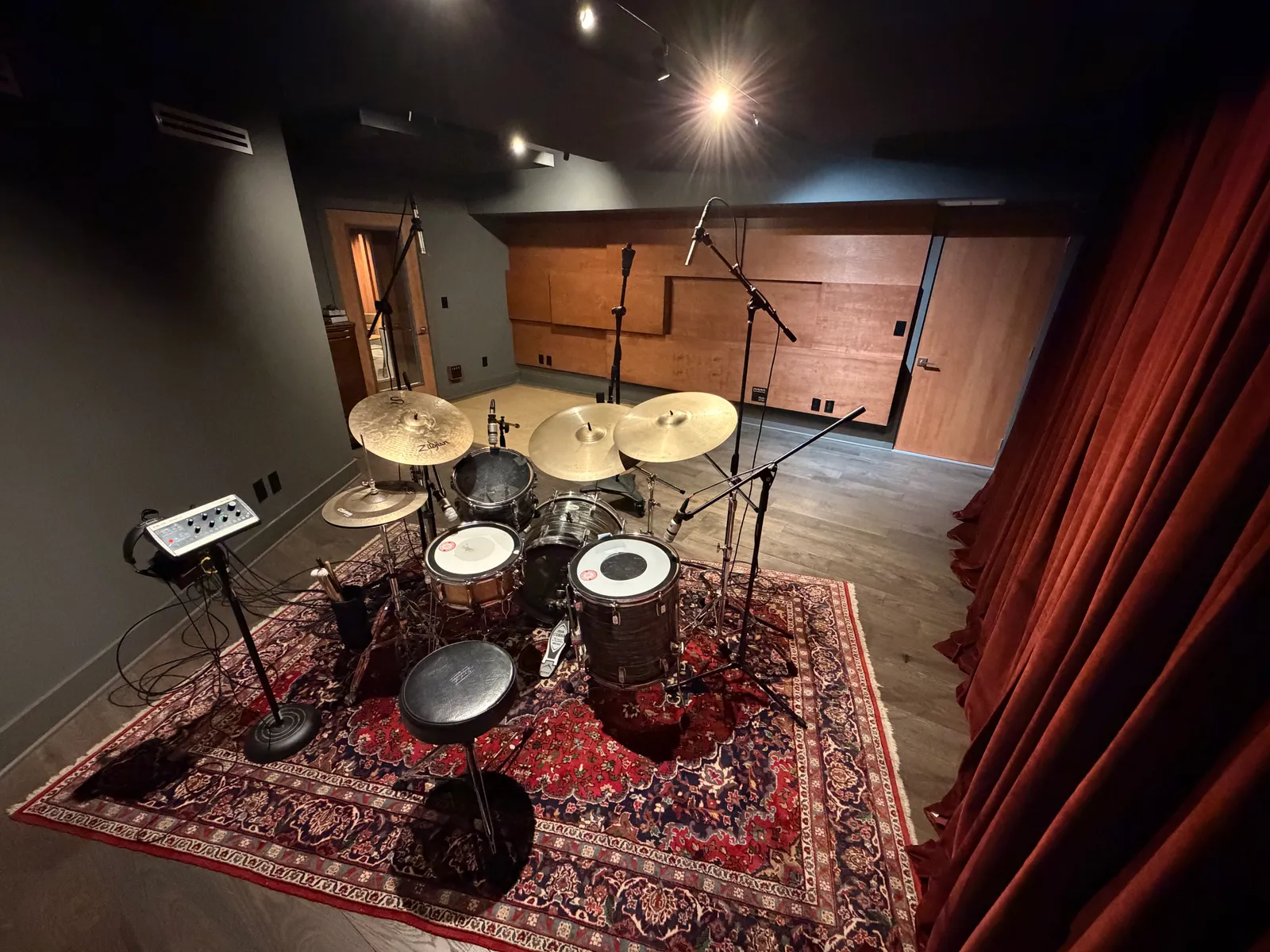 Live room from above. Miked Rogers kit on a Persian rug, curtains to the right, custom wood acoustic panels on far wall. Street-level load-in door to the left.