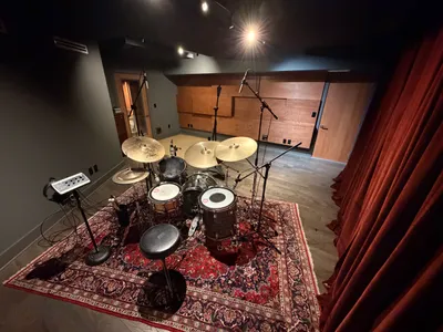 Live room from above. Miked Rogers kit on a Persian rug, curtains to the right, custom wood acoustic panels on far wall. Street-level load-in door to the left.